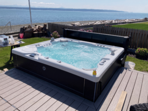 A HotSpring Grandee hot tub is seen on a customer's property on Camano Island, Washington. The large hot tub has a deck surrounding two sides and a fence and beach in the background.