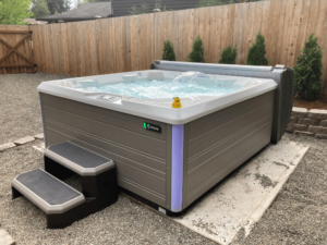 A Hot Spring Flash hot tub viewed from an angle, with two steps on a concrete pad, surrounded by gravel, in a customer's yard in Kenmore.